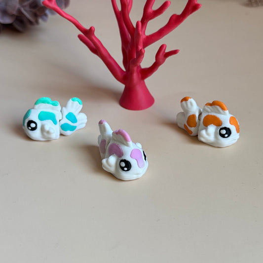 Three mini 3d printed koi fish with colourful markings on a beige surface, with a pink coral-like structure in the background.