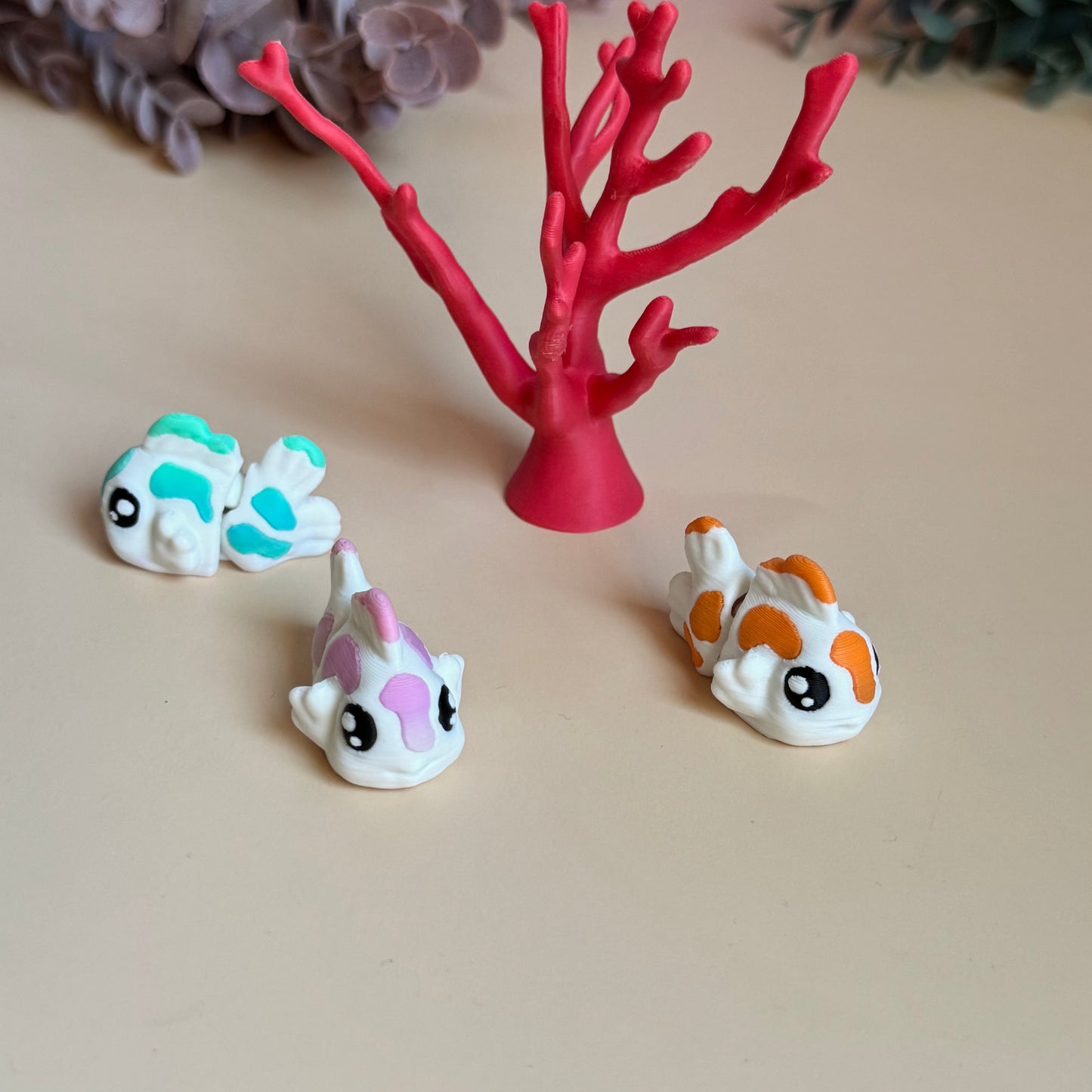 Three mini 3d printed koi fish with colourful markings on a beige surface, with a pink coral-like structure in the background.