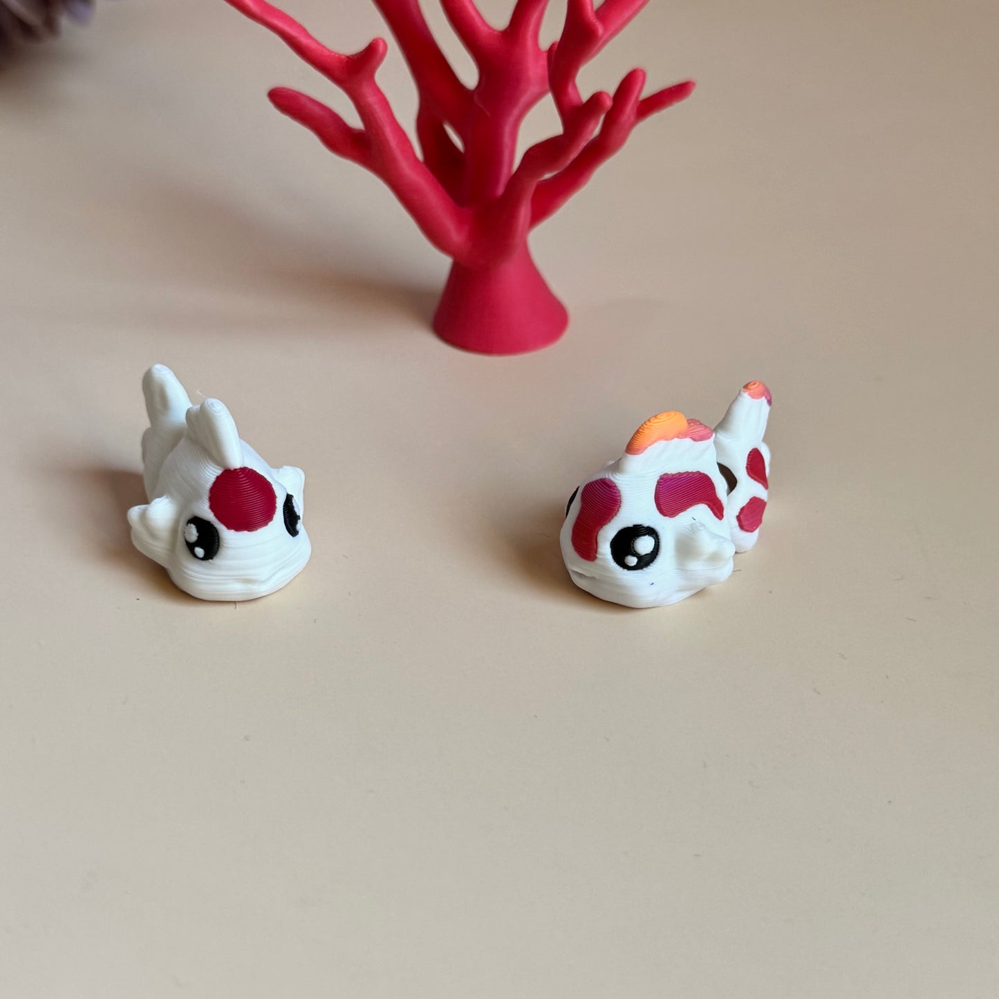 Two mini 3d printed koi fish with colourful markings on a beige surface, with a pink coral-like structure in the background.