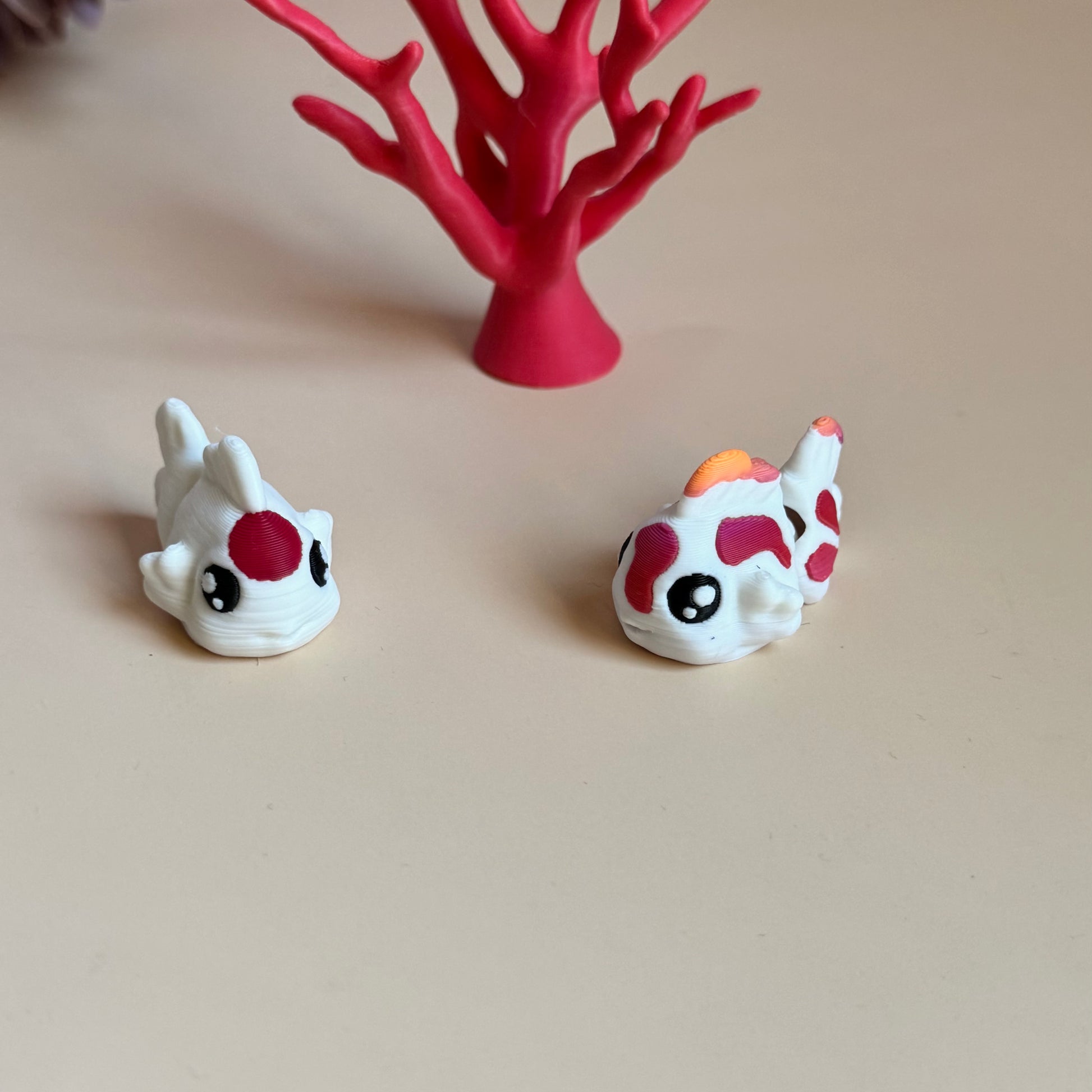Two mini 3d printed koi fish with colourful markings on a beige surface, with a pink coral-like structure in the background.