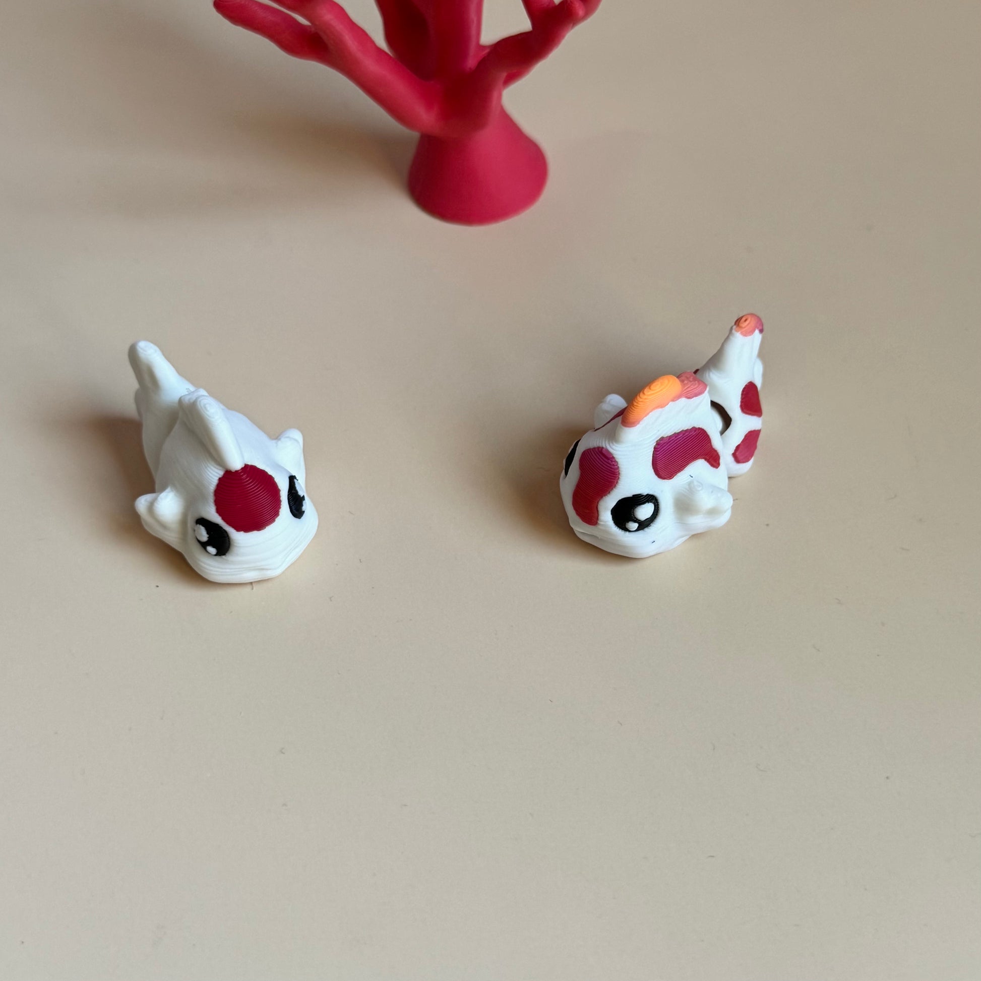 Two mini 3d printed koi fish with colourful markings on a beige surface, with a pink coral-like structure in the background.